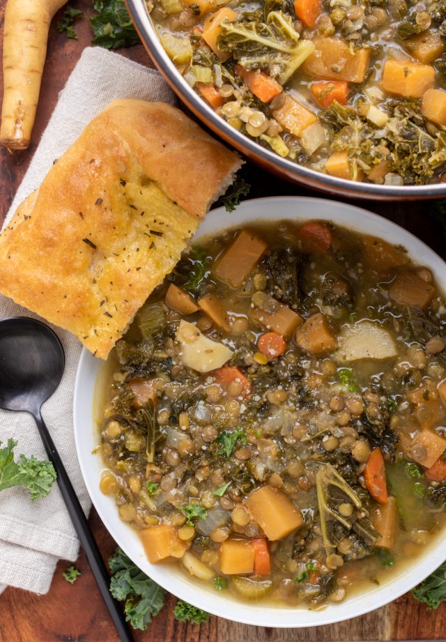 A wooden board with a white bowl of vegetable lentil soup. There's a piece of golden crusty bread next to it, a black round spoon and chopped kale sprinkled on the wooden board. A larger dutch oven pot of soup is in the background.