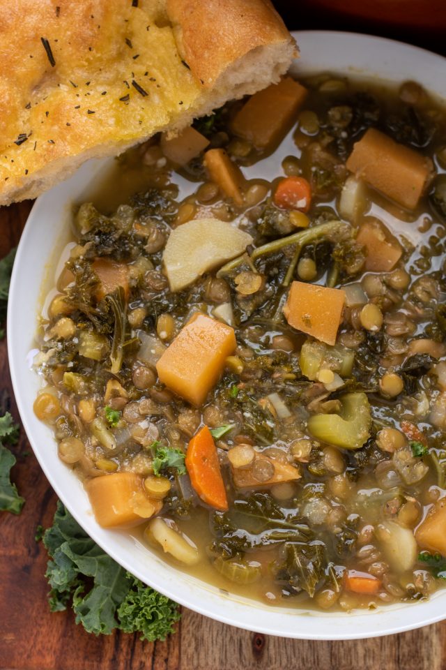 A wooden board with a white bowl full of lentil soup with winter vegetables. There's chopped green kale sprinkled around the bowl with a slice of golden bread. You can see the carrots and turnips in the soup.