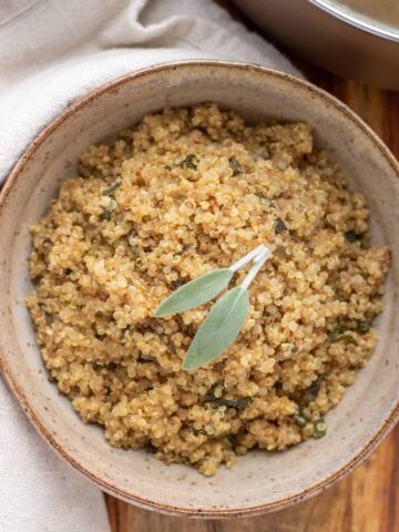 A brown bowl filled with toasted brown butter quinoa that's topped with two fresh sage leaves. It's on top of a wooden surface with a beige cloth next to the bowl.