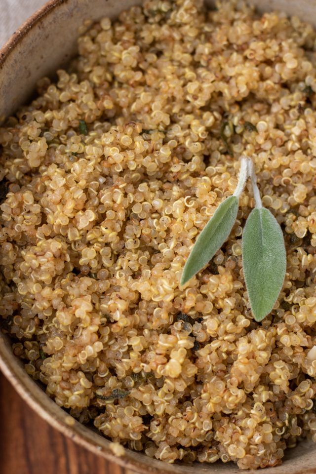 A brown speckled bowl on top of a wood surface. the bowl is filled with quinoa and topped with two fresh sage leaves.