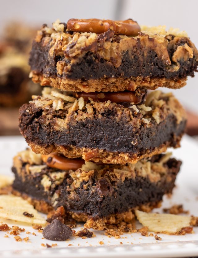 3 brownies stacked on top of each other. The middle is fudgy and they have a pretzel crust with potato chip pretzel topping. There's crushed potato chips and chocolate chips in the background.