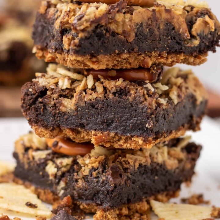 3 brownies stacked on top of each other. The middle is fudgy and they have a pretzel crust with potato chip pretzel topping. There's crushed potato chips and chocolate chips in the background.