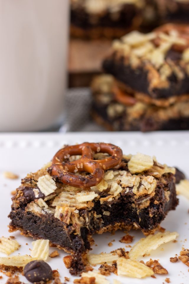 A brownie on a white plate with a bite taken out of it. It's topped with a pretzel and crushed potato chips. There's crumbs on the plate and a glass of milk with more brownies in the background.