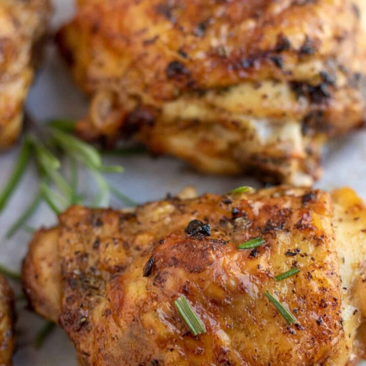 Golden and crispy chicken thighs on white parchment paper. There's a sprinkle of rosemary on top of the chicken with a fresh rosemary sprig in the background with a lemon.