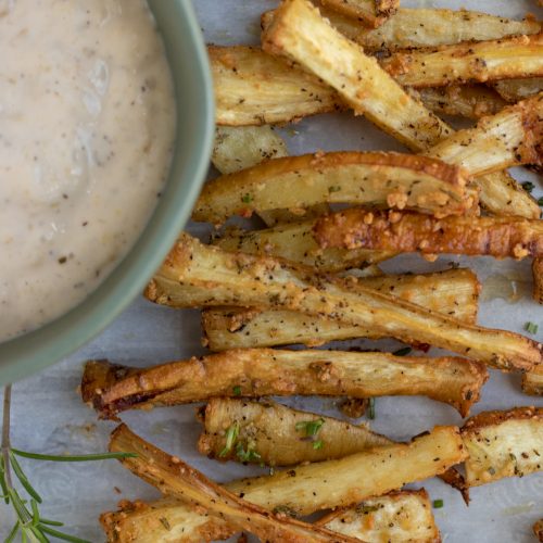 Parsnip fries that are sprinkled with rosemary and parmesan. They're golden and crispy with a small round bowl of dipping sauce next to them.