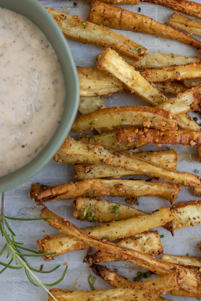 Parsnip fries that are sprinkled with rosemary and parmesan. They're golden and crispy with a small round bowl of dipping sauce next to them.