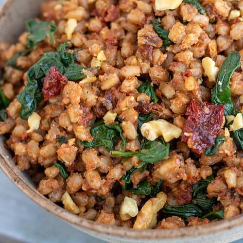 A brown speckled bowl filled with farro covered in sun-dried tomato pesto with fresh spinach and toasted walnuts.