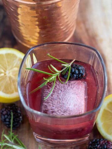 A square rocks glass with a large square ice cube, fresh rosemary sprig and a blackberry. It's filled with blackberry smash cocktail with fresh lemons and a copper cocktail shaker in the background.