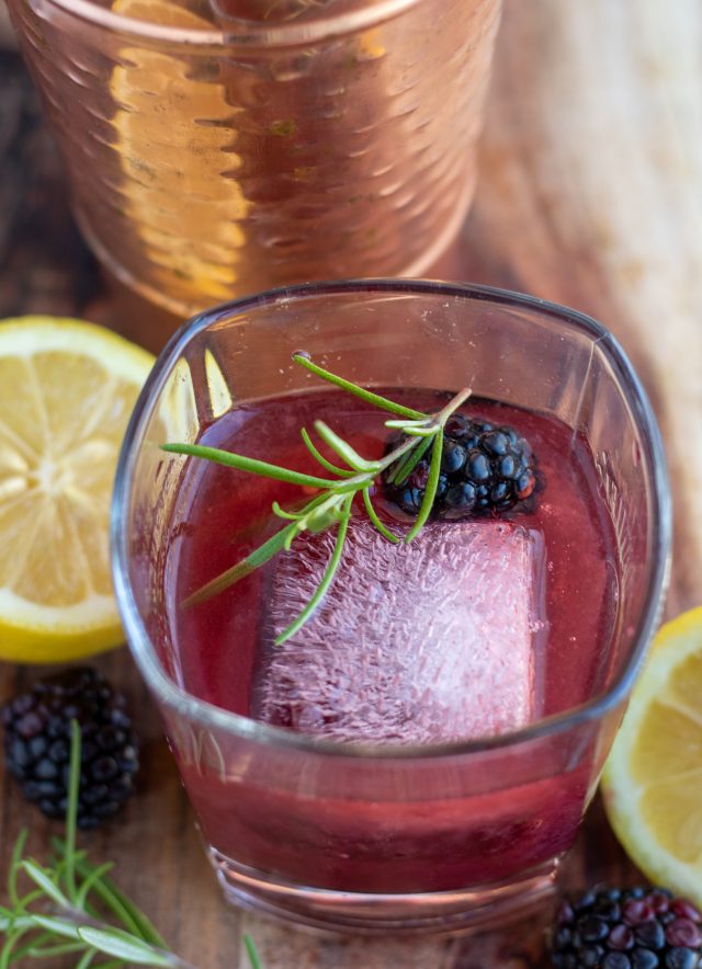 A square rocks glass with a large square ice cube, fresh rosemary sprig and a blackberry.  It's filled with blackberry smash cocktail with fresh lemons and a copper cocktail shaker in the background.