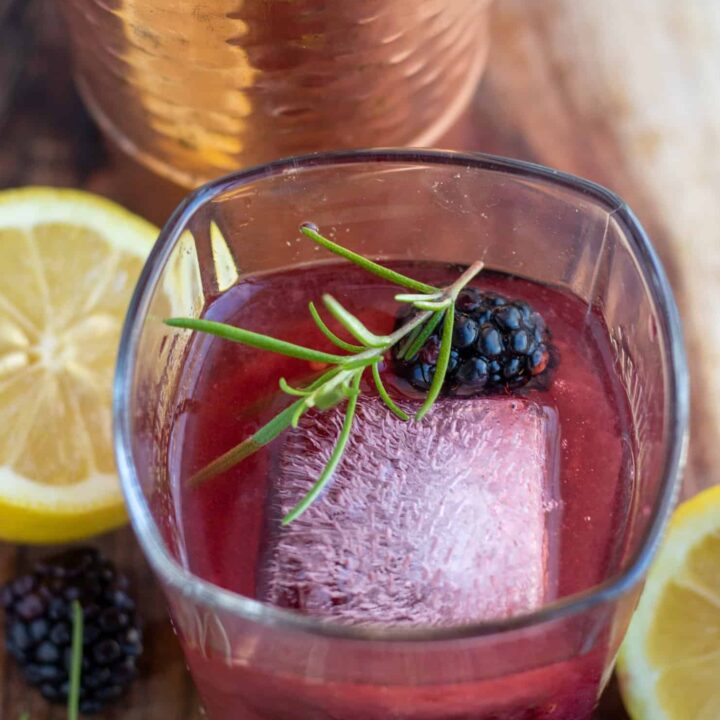 A square rocks glass with a large square ice cube, fresh rosemary sprig and a blackberry. It's filled with blackberry smash cocktail with fresh lemons and a copper cocktail shaker in the background.