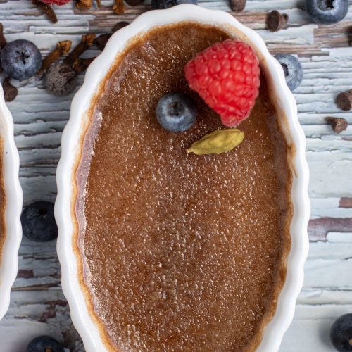 A white oval ramekin filled with chocolate chai creme brulee that's topped with fresh berries. There's fresh blueberries, raspberries, whole cardamom pods and chocoalte chips in the background.