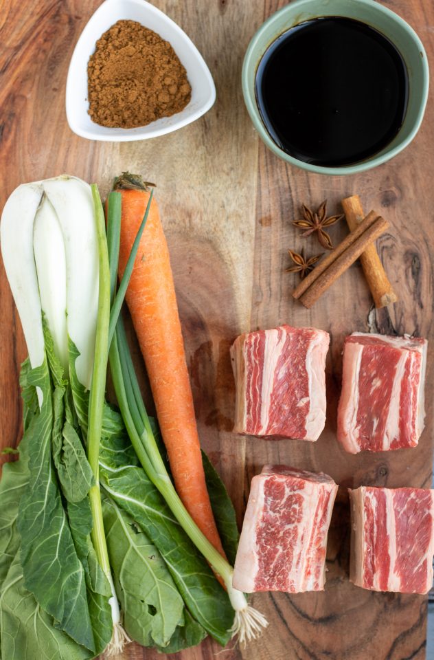 A wooden cutting board topped with 4 raw pieces of beef short ribs, 1 bunch of bok choy, a whole carrot, cinnamon stick and star anise. There's two small bowls in the background; one with chinese five spice, one with soy sauce