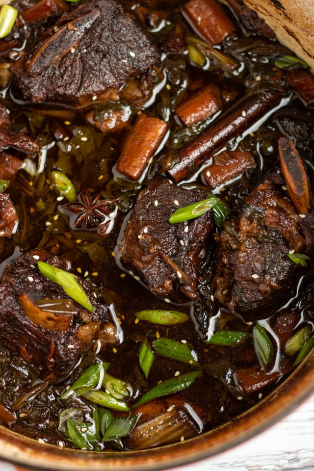 A dutch oven filled with braised asian short ribs. The sauce is dark brown with carrots and bright green scallions. There's sesame seeds sprinkled over the top