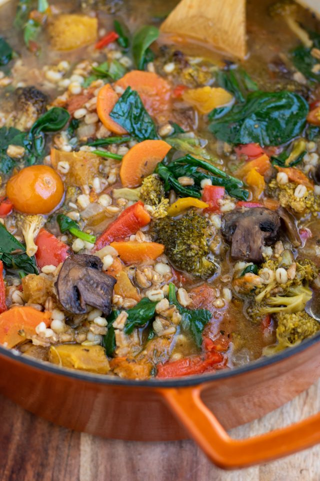 An orange dutch oven pan on top of a wooden surface filled with color vegetable barley soup. You can see the orange tomaotes, mushrooms and broccoli