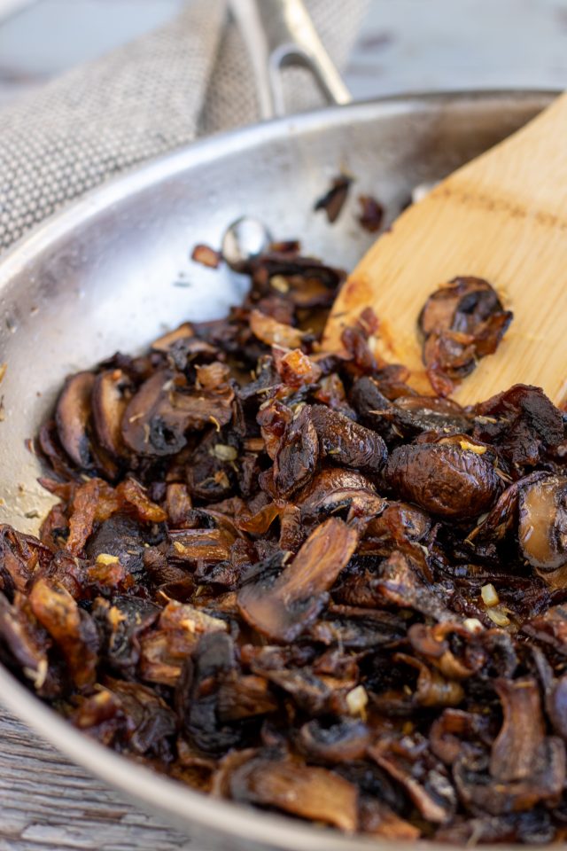 A stainless steel saute pan with caramelized mushrooms and onions with a wooden spoon scooping them up. The mushrooms are a deep brown and slightly crispy on the edges