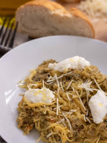 A white pasta bowl with spaghetti squash topped with broccoli pesto and dollops of ricotta cheese. It's drizzled with olive oil and grated parmesan cheese. There's a fork next to the bowl and a slice of Italian bread in the background
