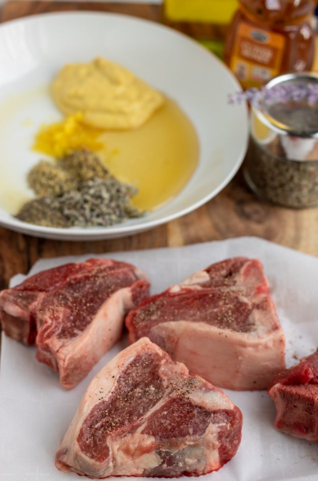 A wooden cutting board with raw lamb loin chops on it. There's a white bowl of the marinade ingredients and a jar of honey in the background.