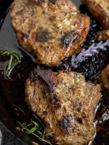 A cast iron pan with seared lamb chops baked in it. They're golden brown with fresh rosemary and lavender in the pan.