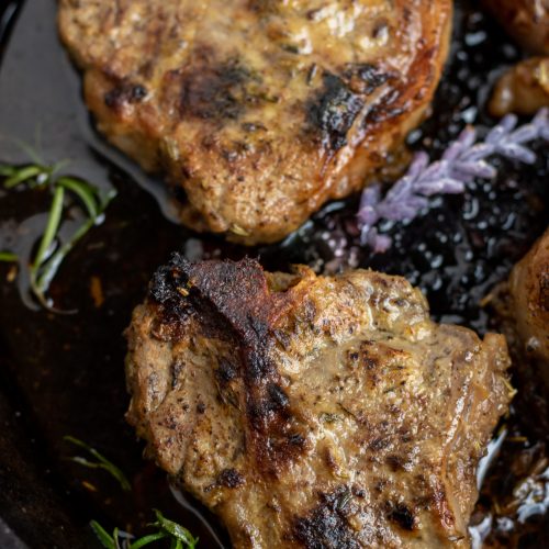 A cast iron pan with seared lamb chops baked in it. They're golden brown with fresh rosemary and lavender in the pan.