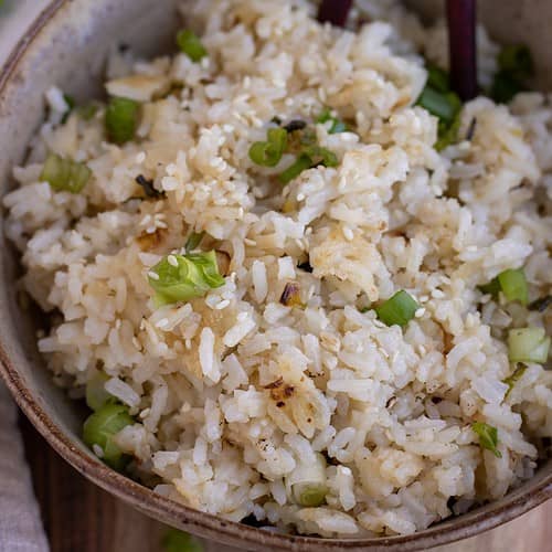 A brown speckled bowl filled with white rice that's slightly crispy and has scallions on top and sesame seeds. There's a pair of chopsticks sticking out of the bowl. There's a beige cloth napkin next to the bowl on a wooden surface with sliced scallions and white rice in the background