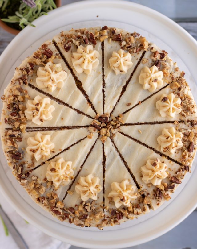 An overhead shot of a decorated carrot cake on a white cake stand. It's got crushed nuts sprinkled around the border and it's sliced into 12 equal slices. There's a lavender plant in the background.