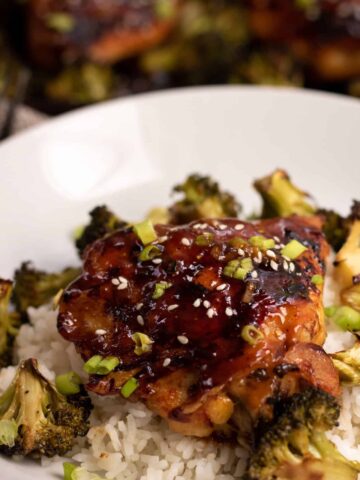 A shallow white bowl with white rice, roasted broccoli and a piece of crispy chicken thighs that have an Asian sticky glaze on them. The chicken is browned and sprinkled with sesame seeds and scallions. There's a sheet pan of chicken in the background.
