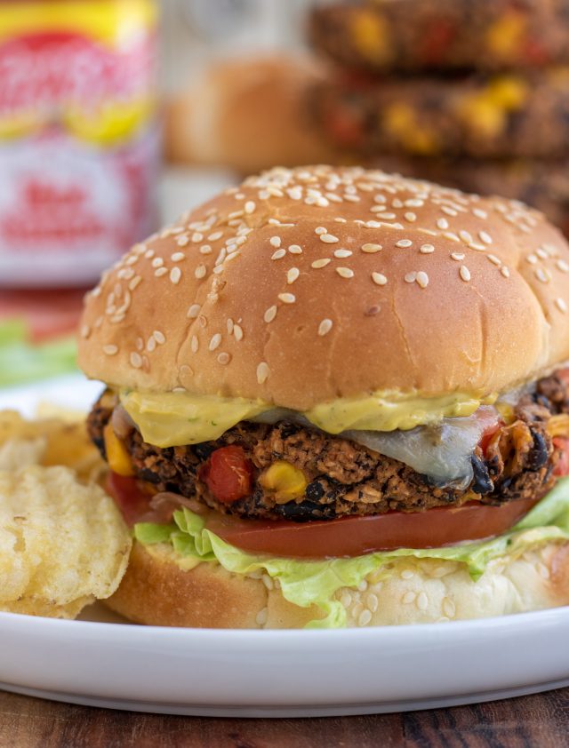 A round white plate with a black bean burger on a sesame seed bun with lettuce, tomato and sauce dripping down the side. There's ruffled potato chips on the plate, a stack of burgers in the background and a bottle of hot sauce