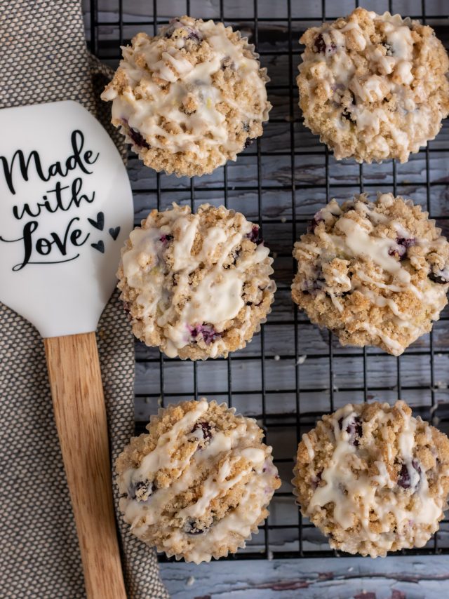 A cooling rack topped with six blueberry streusel muffins. There's a spatula next to the muffins that says "Made with Love"