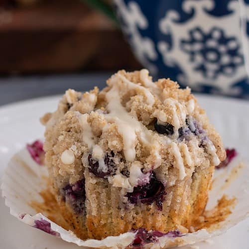 A small white plate topped with a blueberry muffin. The liner has been peeled off the muffin and it's topped with crumble topping with glaze dripping down it. You can see more muffins in the background on a wooden board and a blue and white swirled tea cup with a tea bag hanging out of it