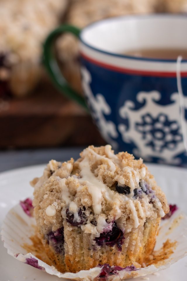 A small white plate topped with a blueberry muffin. The liner has been peeled off the muffin and it's topped with crumble topping with glaze dripping down it. You can see more muffins in the background on a wooden board and a blue and white swirled tea cup with a tea bag hanging out of it