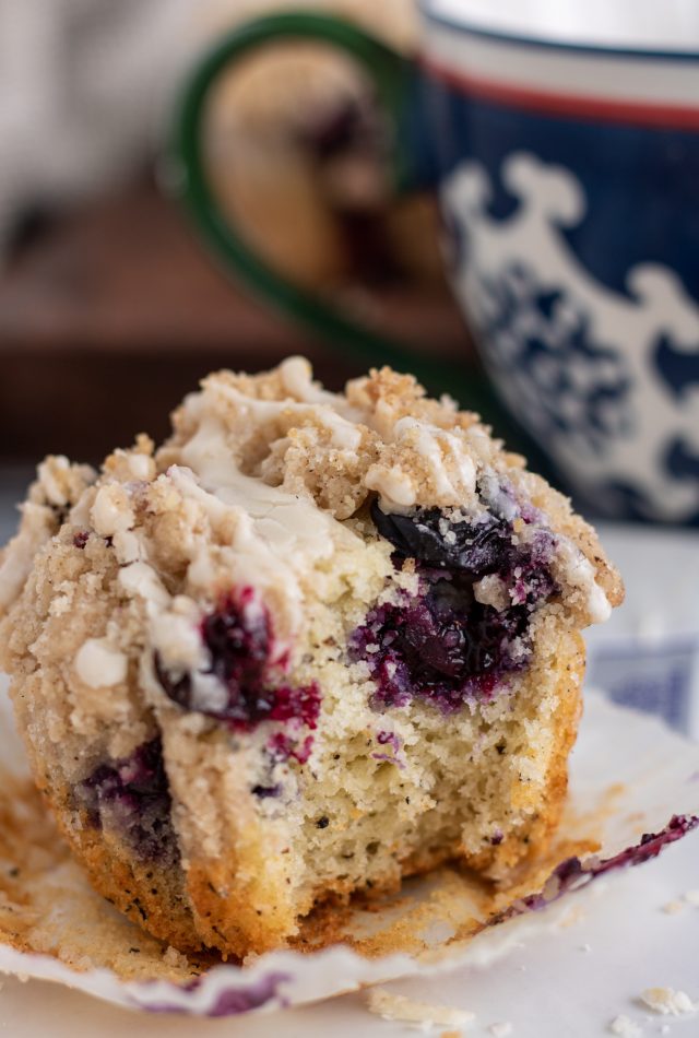 A small white plate with a Earl Grey blueberry muffin on it. The liner has been peeled back and the muffin has a bite taken out of it. There's a blue and white swirl tea cup in the background