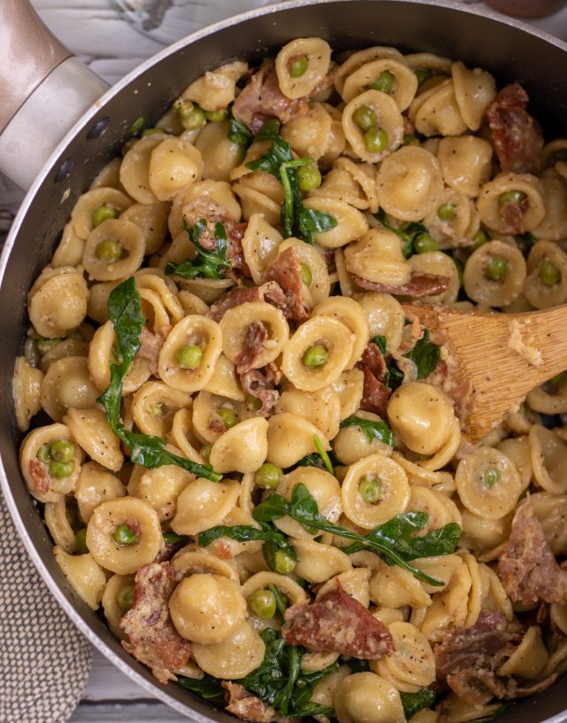 A large bronze sauce pan filled with creamy orecchiette pasta tossed with peas, crispy prosciutto and arugula.