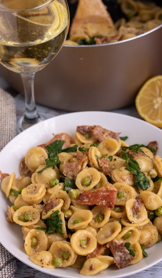 A white pasta bowl filled with orecchiette pasta, peas and prosciutto. There's a gold large saucepan in the background with more pasta and a glass of wine in the background.