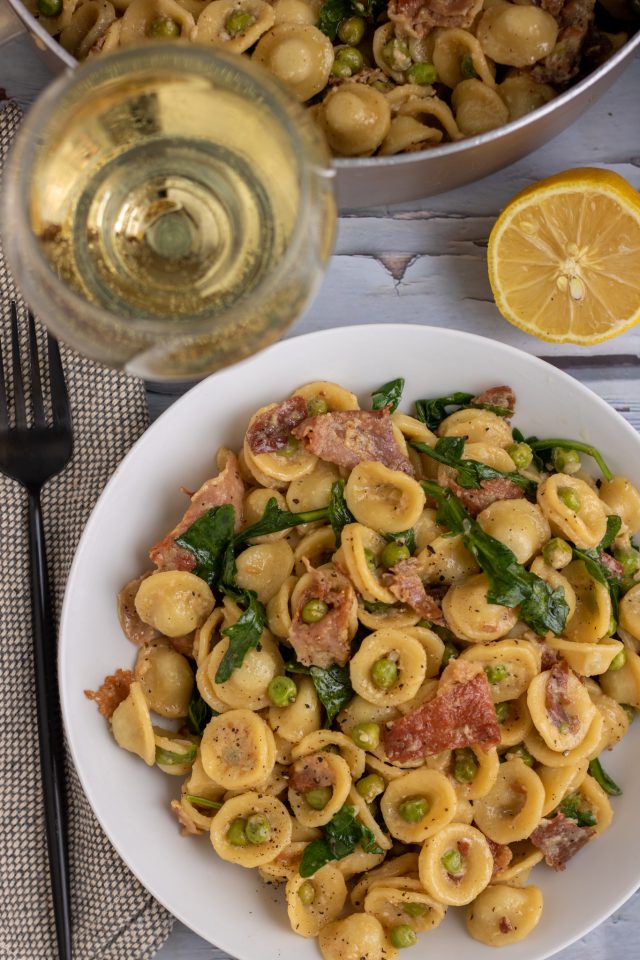 A white pasta bowl filled with creamy lemon pasta with peas, prosciutto and arugula. There's a glass of white wine next to the plate with a half a lemon and a large pot of pasta in the background