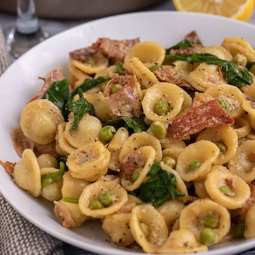 A white pasta bowl filled with orecchiette pasta with peas, crispy prosciutto and arugula. There's a glass of white wine and half a lemon in the background.