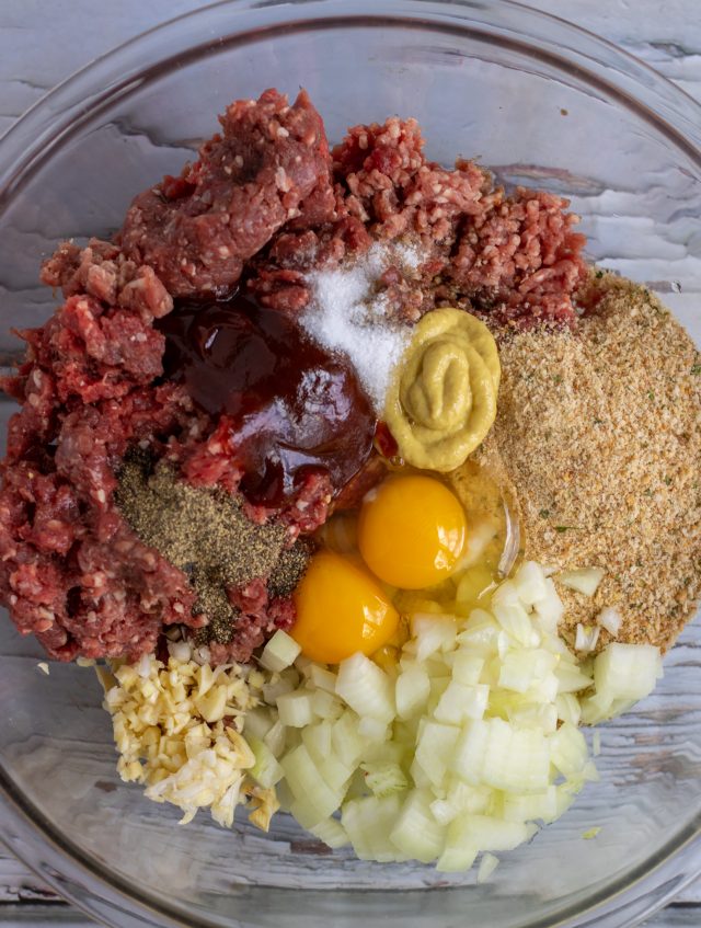 A large glass bowl of ground beef, breadcrumb, egg, BBQ sauce, dijon mustard, onions and garlic.