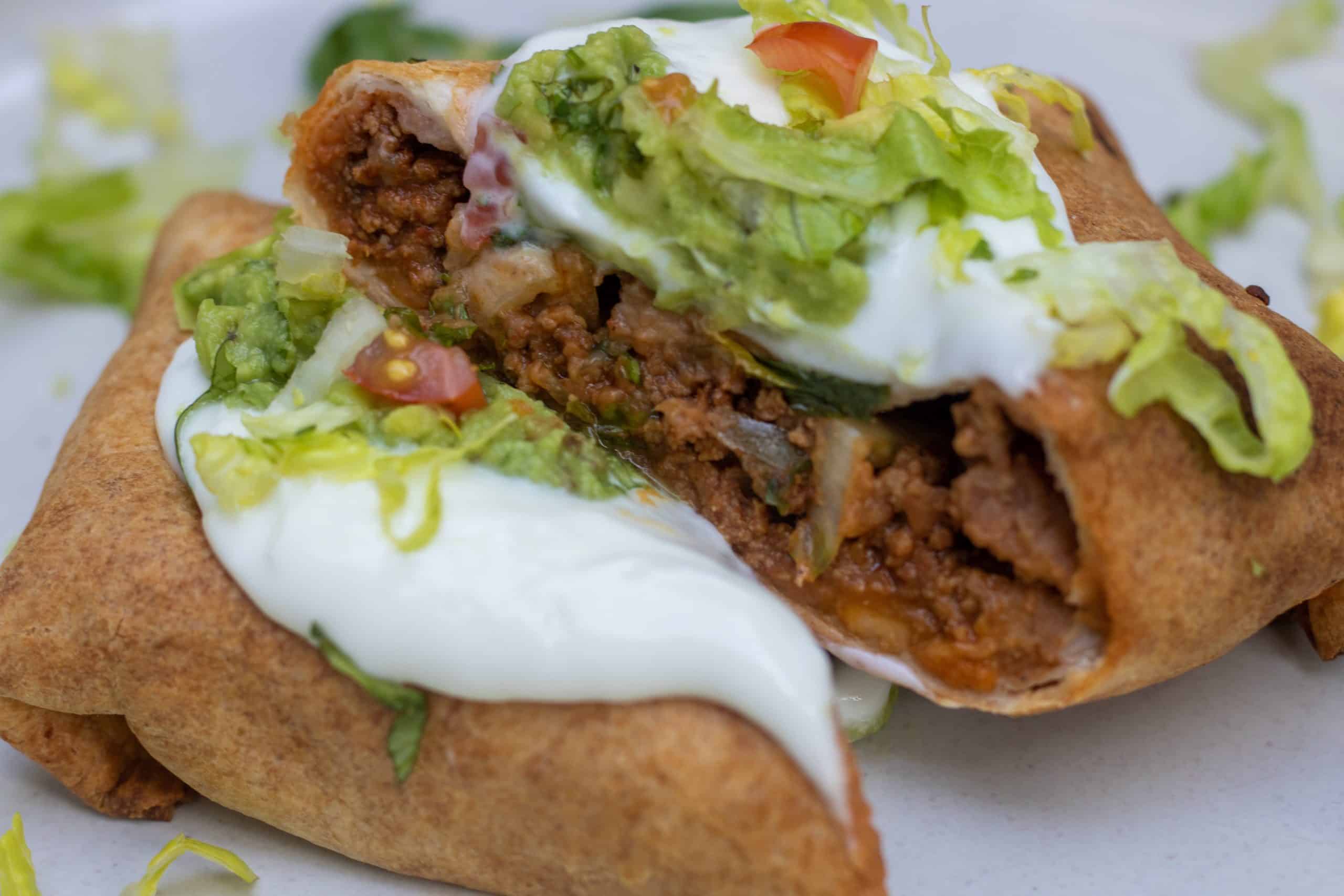 A crispy healthy air fryer beef chimichanga that's cut in half so you can see the ground beef and refried bean filling. It's topped with sour cream, guacamole and shredded lettuce