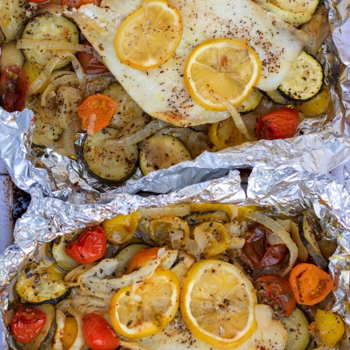 Two foil packets with white fish, colorful cherry tomatoes, sliced zucchini and sliced onions. It’s sprinkled with salt and pepper and there are slices of lemon on top of the fish.