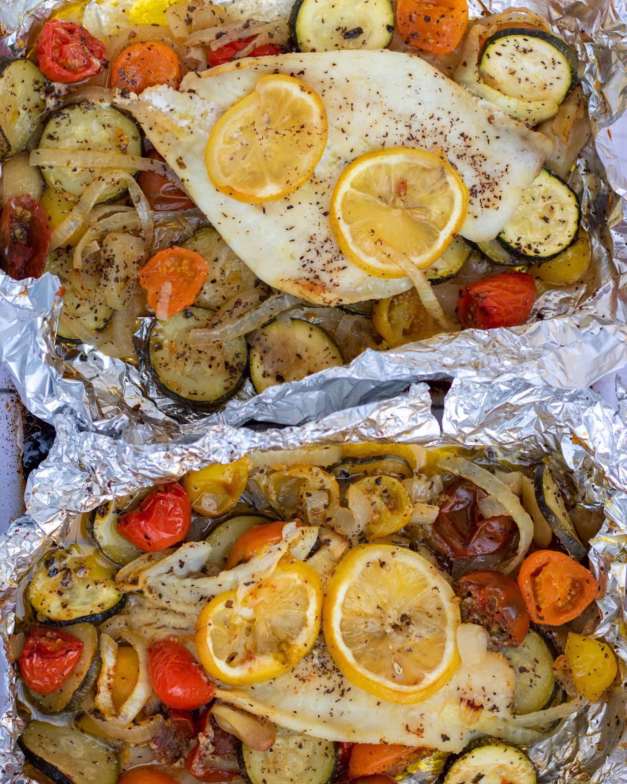 Two foil packets with white fish, colorful cherry tomatoes, sliced zucchini and sliced onions. It’s sprinkled with salt and pepper and there are slices of lemon on top of the fish.