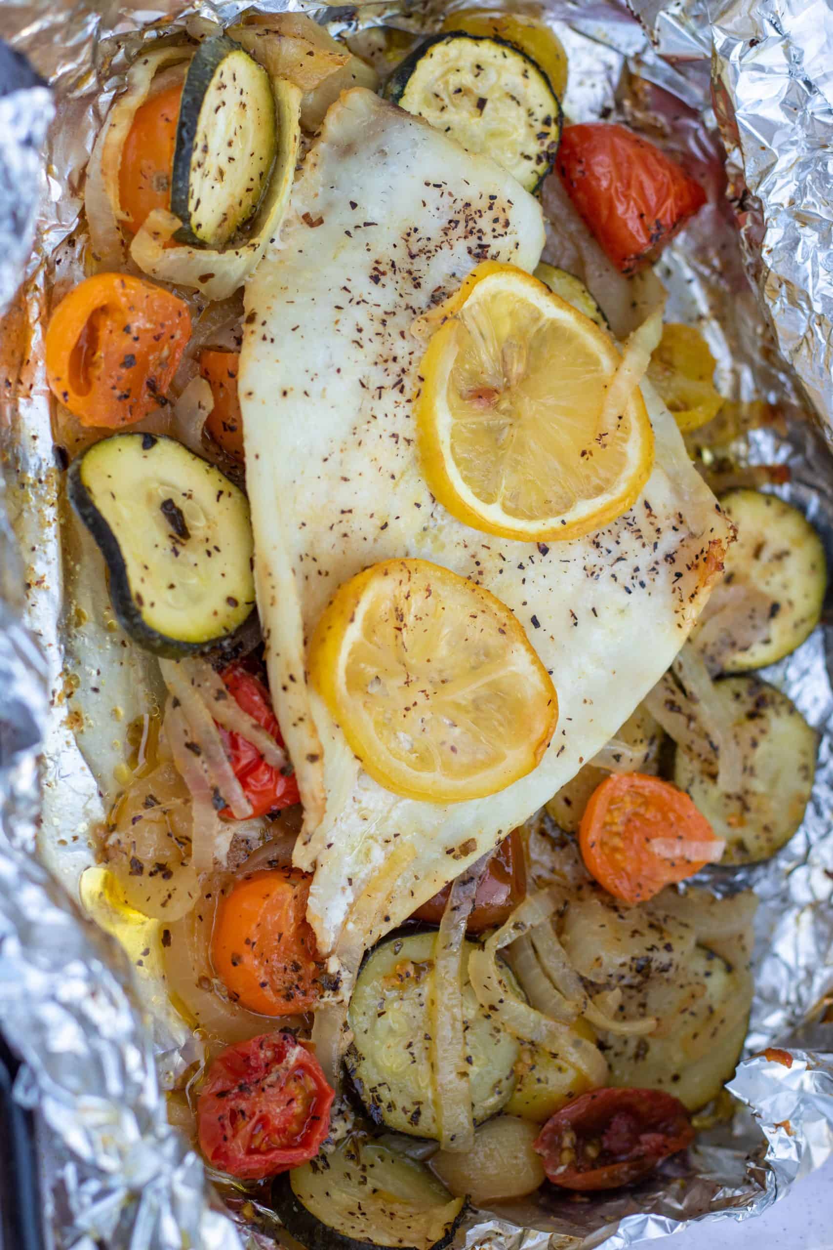 A single piece of white fish in foil, surrounded by roasted cherry tomatoes that are yellow, red and orange, sliced zucchini and caramelized onions. The fish is baked in the oven in a foil packet. Two slices of lemon are on top of the fish