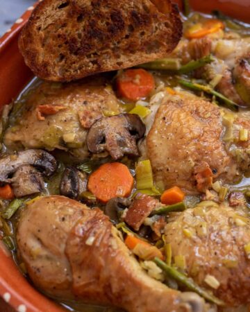 A round rustic casserole dish with coq au vin blanc with carrots, mushrooms, asparagus and leeks. There's a piece of crusty golden bread on the edge of the dish and the chicken skin is golden brown