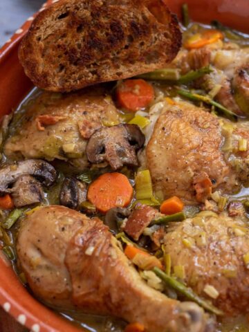 A round rustic casserole dish with coq au vin blanc with carrots, mushrooms, asparagus and leeks. There's a piece of crusty golden bread on the edge of the dish and the chicken skin is golden brown