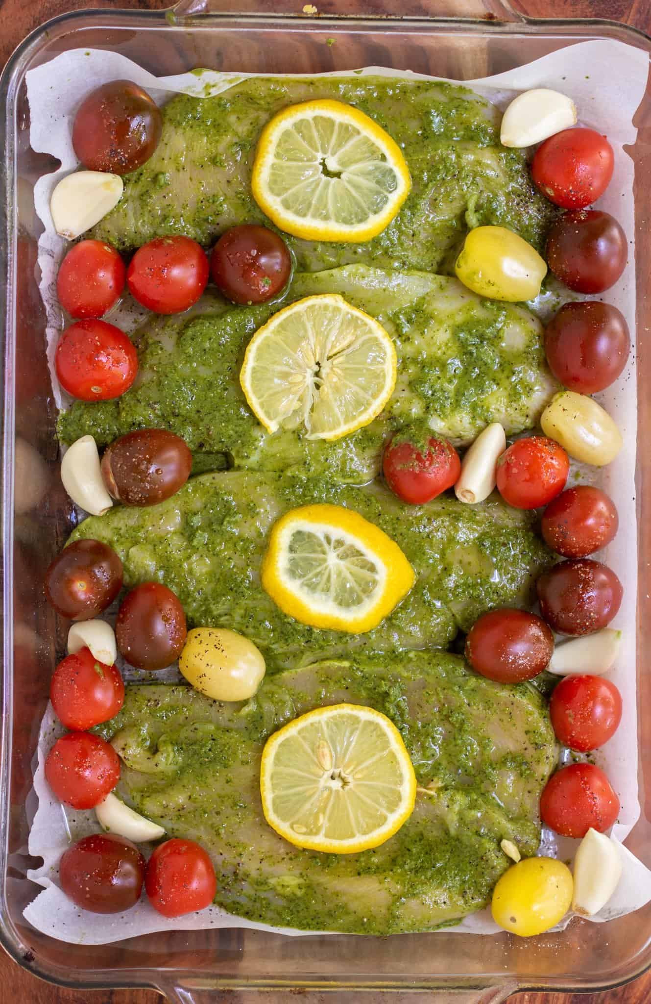 A rectangle glass baking dish lined with parchment with mixed cherry tomaotes, garlic cloves, and pesto marinated chicken breasts that are topped with a lemon slice