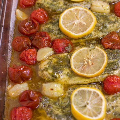 A glass baking dish with four pieces of chicken breasts that are covered in basil pesto and topped with a slice of lemon. There's roasted whole cherry tomatoes and garlic cloves around the chicken.