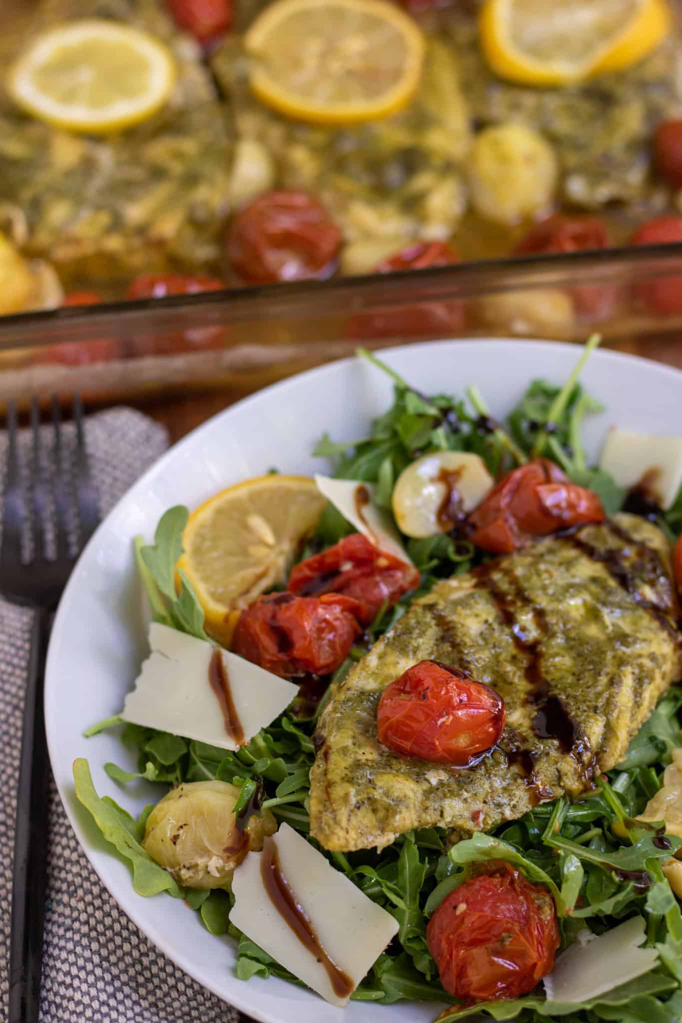 A white pasta dish filled with arugula and topped with pesto chicken. There's shaved Parmesan cheese and a fresh lemon wedge on the dish. It's drizzled with balsamic glaze. You can see a glass baking dish filled with chicken and tomatoes in the background.