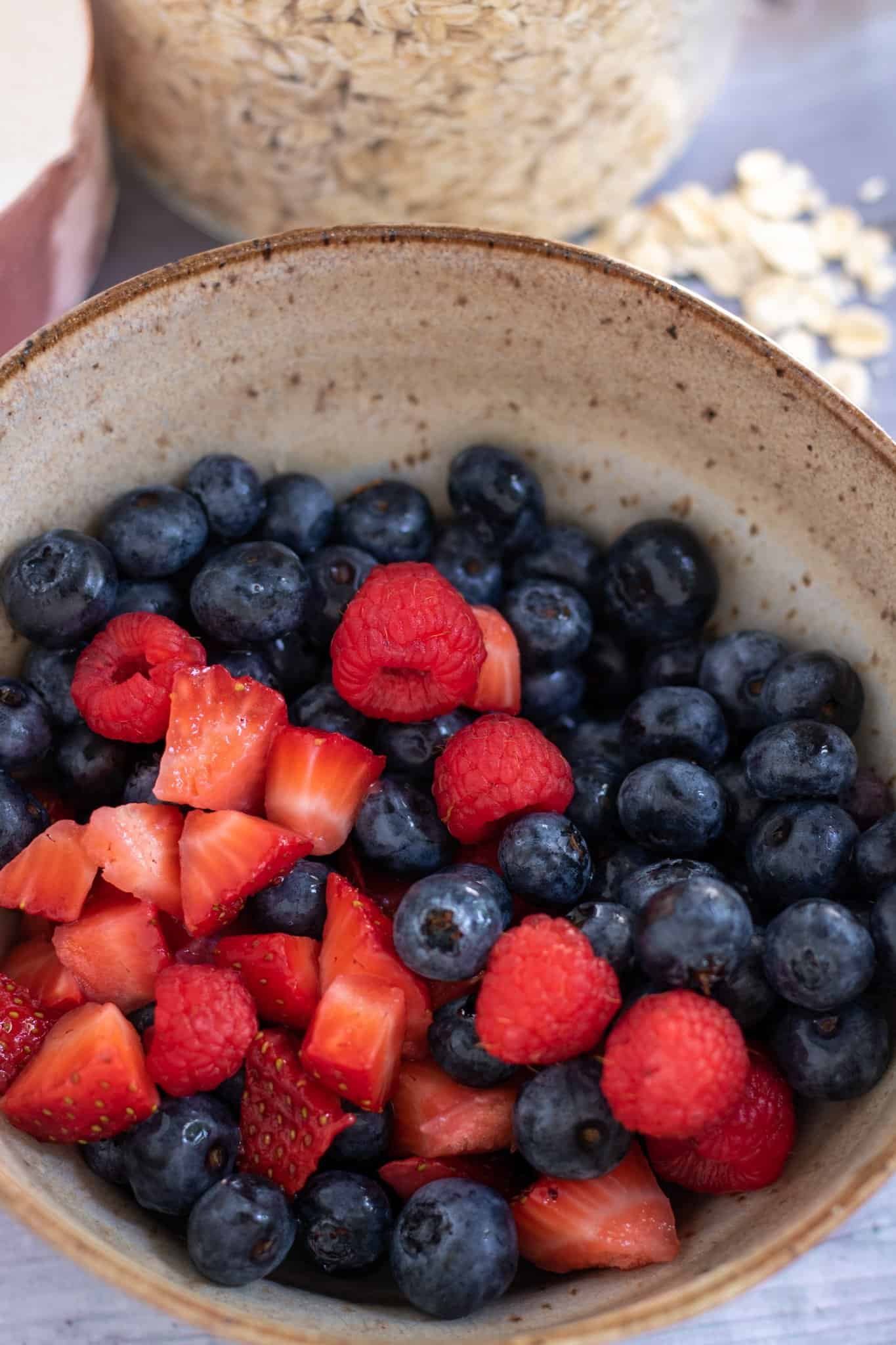 A brown speckled bowl filled with fresh blueberries, raspberries, and strawberries. There's oats sprinkled in the background.