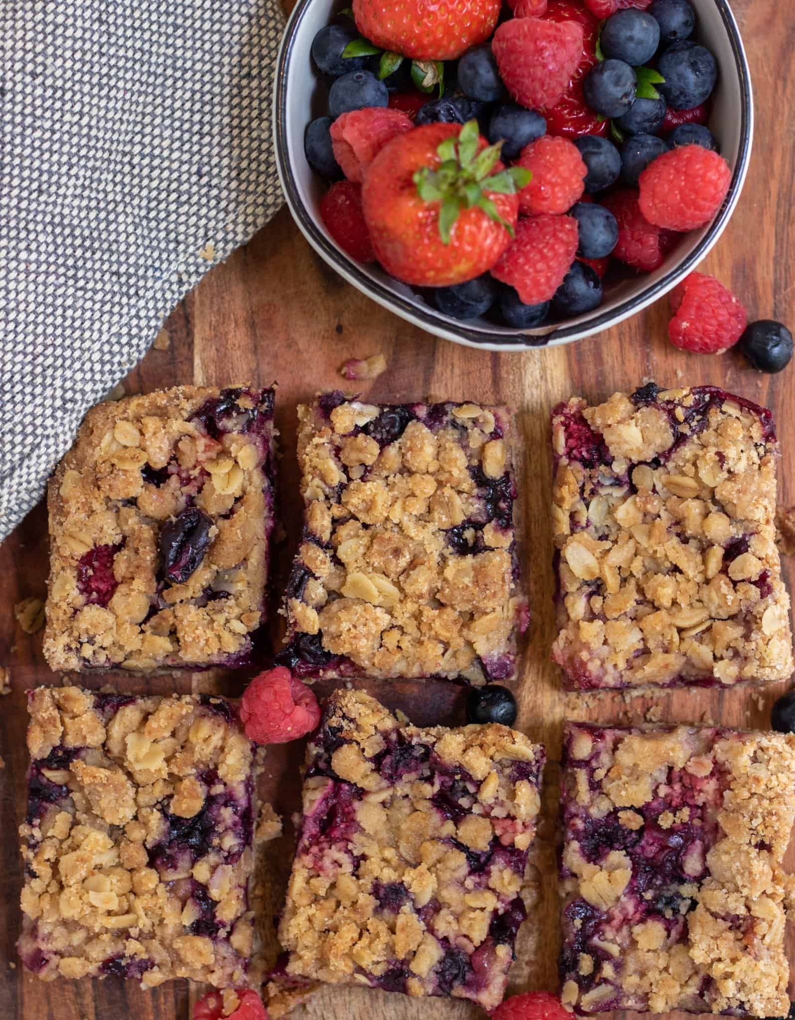 6 square berry crumble bars with a small bowl of fresh mixed berries next to them.