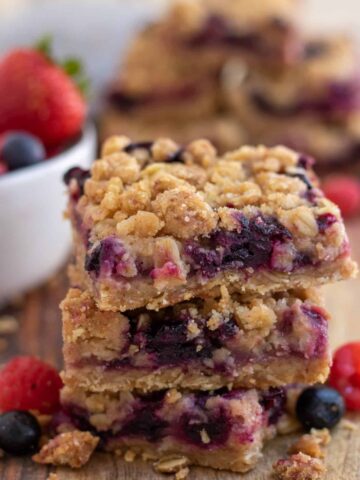 A wooden surface topped with 3 stacked mixed berry oatmeal crumble bars with blueberries and raspberries sprinkled around them.