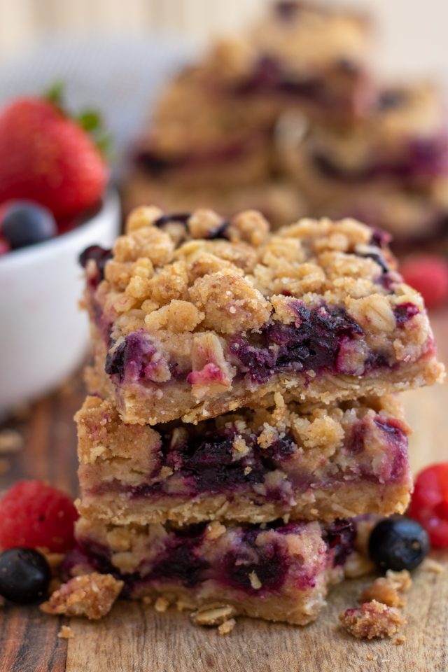 A wooden surface topped with 3 stacked mixed berry oatmeal crumble bars with blueberries and raspberries sprinkled around them.