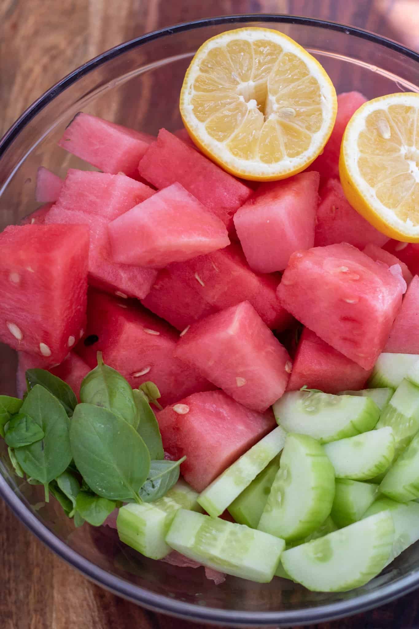 A large glass bowl filled with cubed watermelon, sliced cucumbers, fresh basil leavevs and lemons.